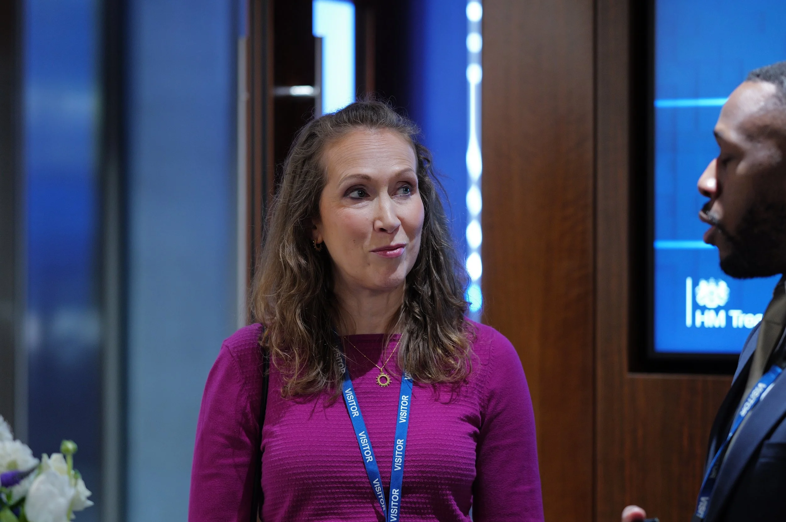 Woman in purple sweater with visitor badge talking to a man in a suit at an indoor event.