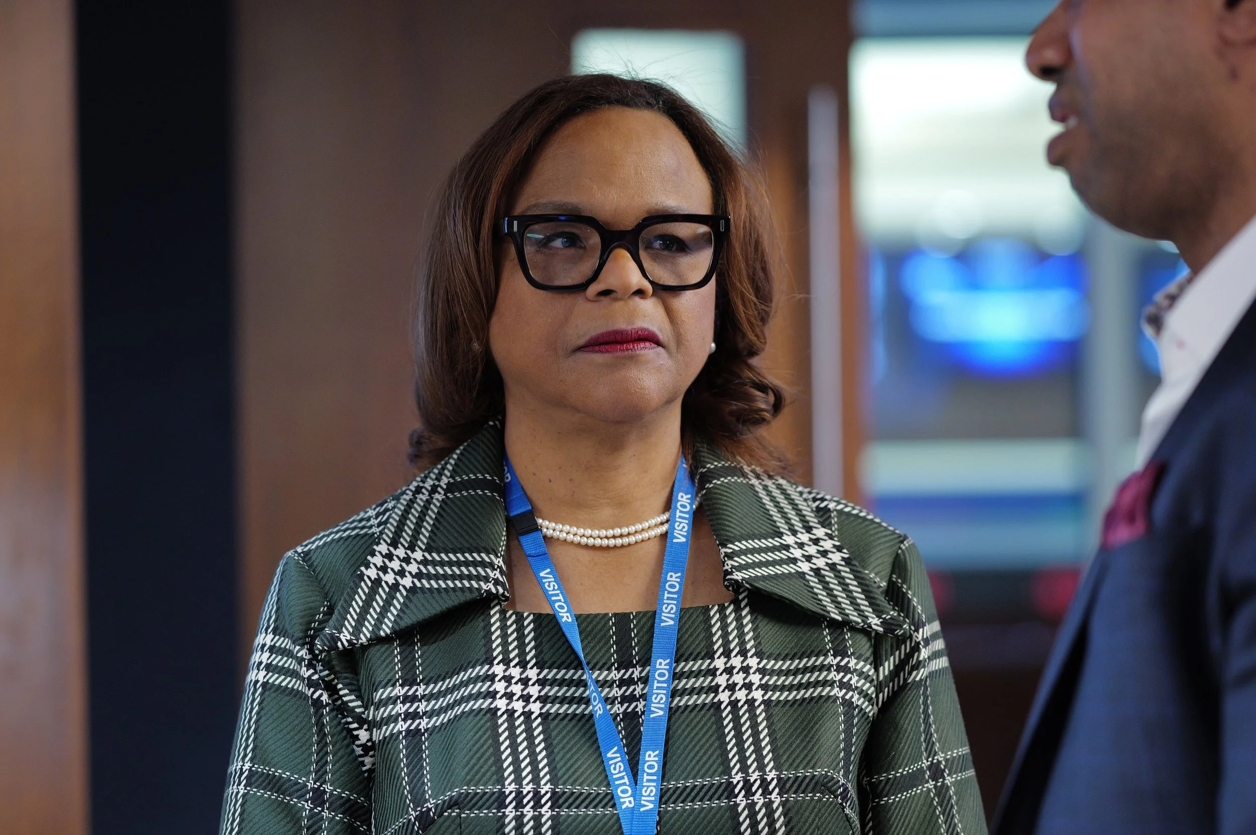 Woman wearing glasses, a green plaid jacket, pearl necklace, and a blue visitor lanyard listening seriously to a man in a suit.