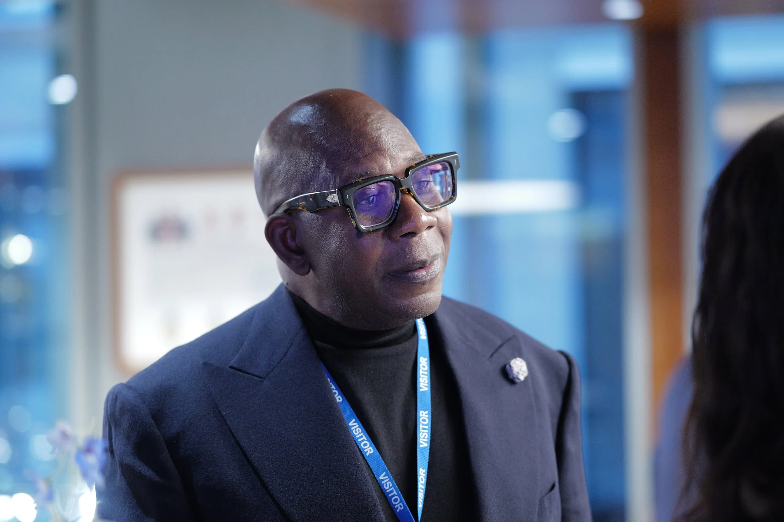 Man wearing glasses and a visitor badge, dressed in a dark blazer and black turtleneck, looking attentively at someone.
