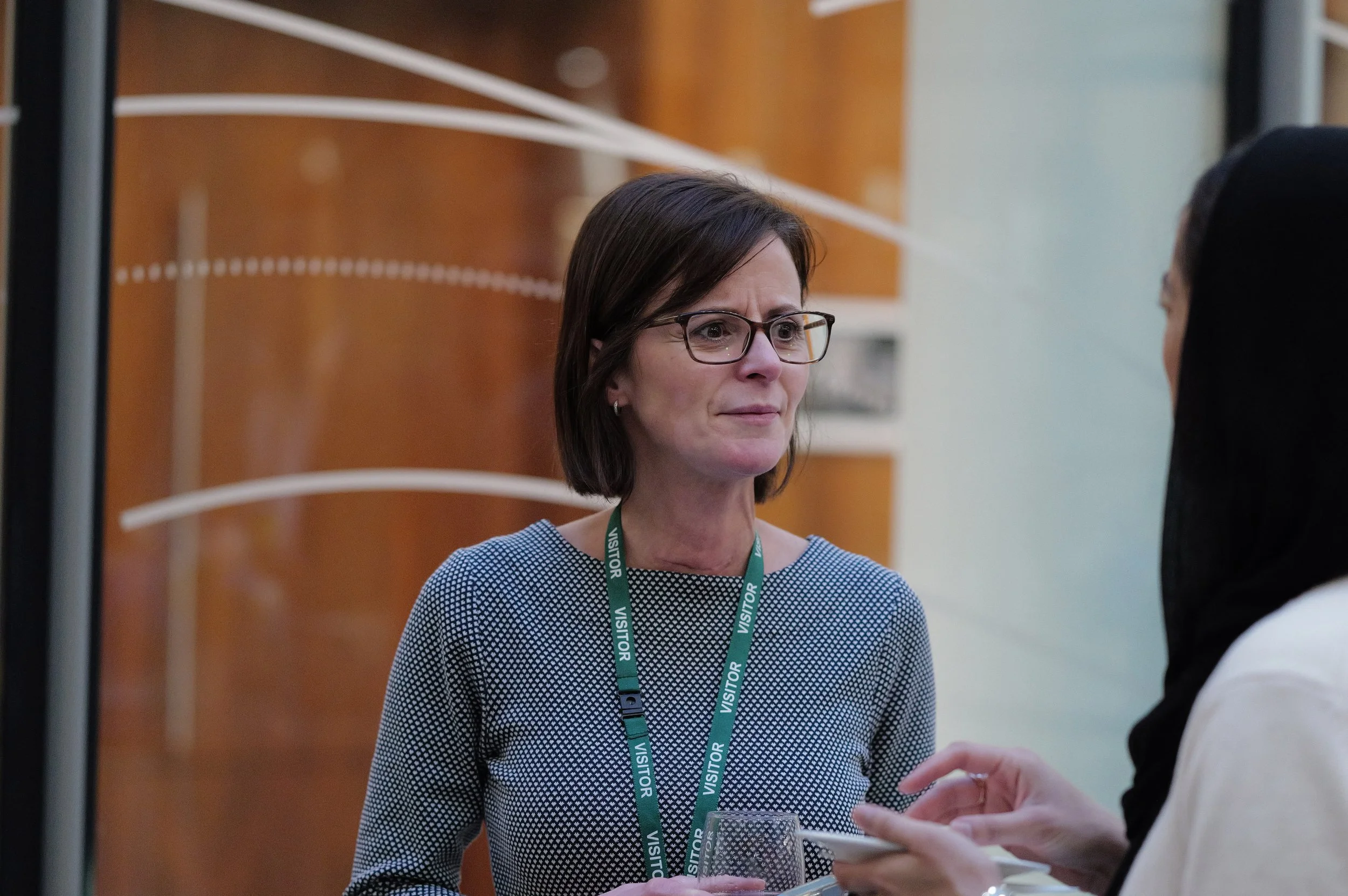 Woman with glasses and a visitor badge talking to another person holding a cup and saucer.