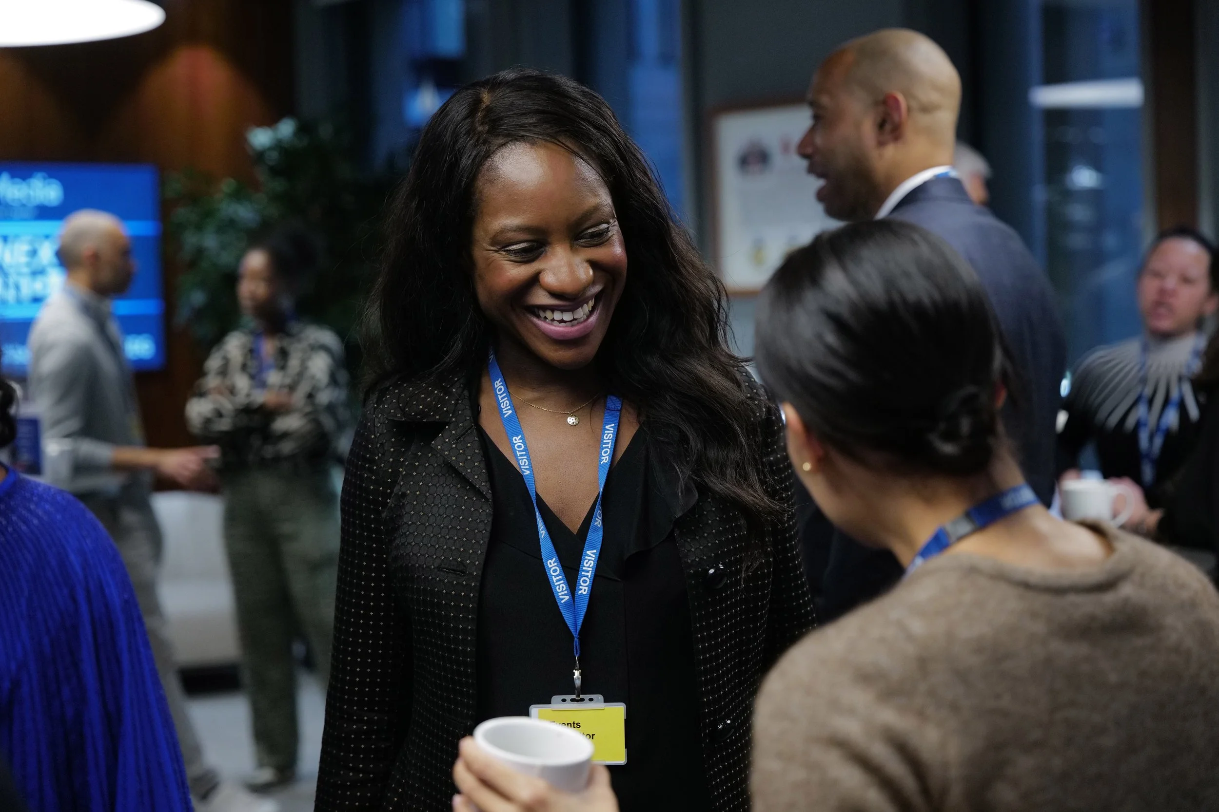 Smiling woman wearing a visitor badge converses with another person holding a coffee cup at a professional event.