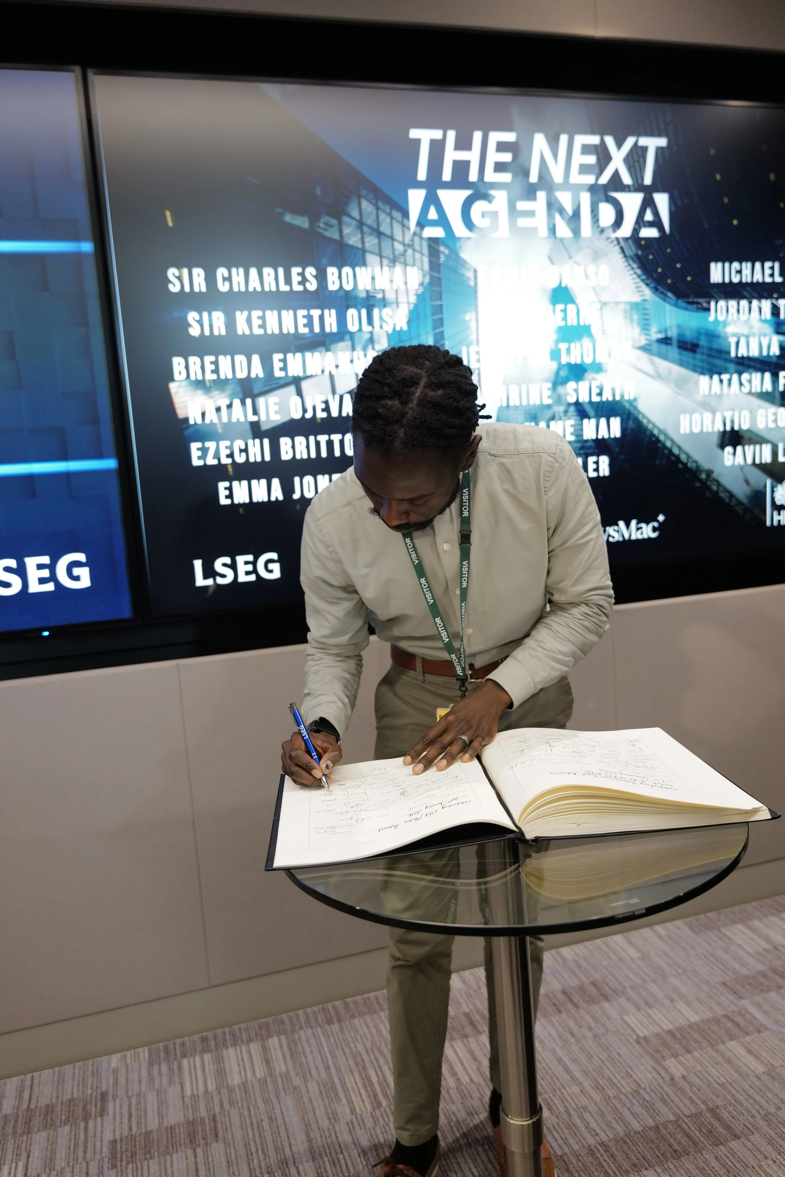 Man in light shirt signing a large guestbook on a glass table in front of a digital display titled 'The Next Agenda' with multiple names listed.