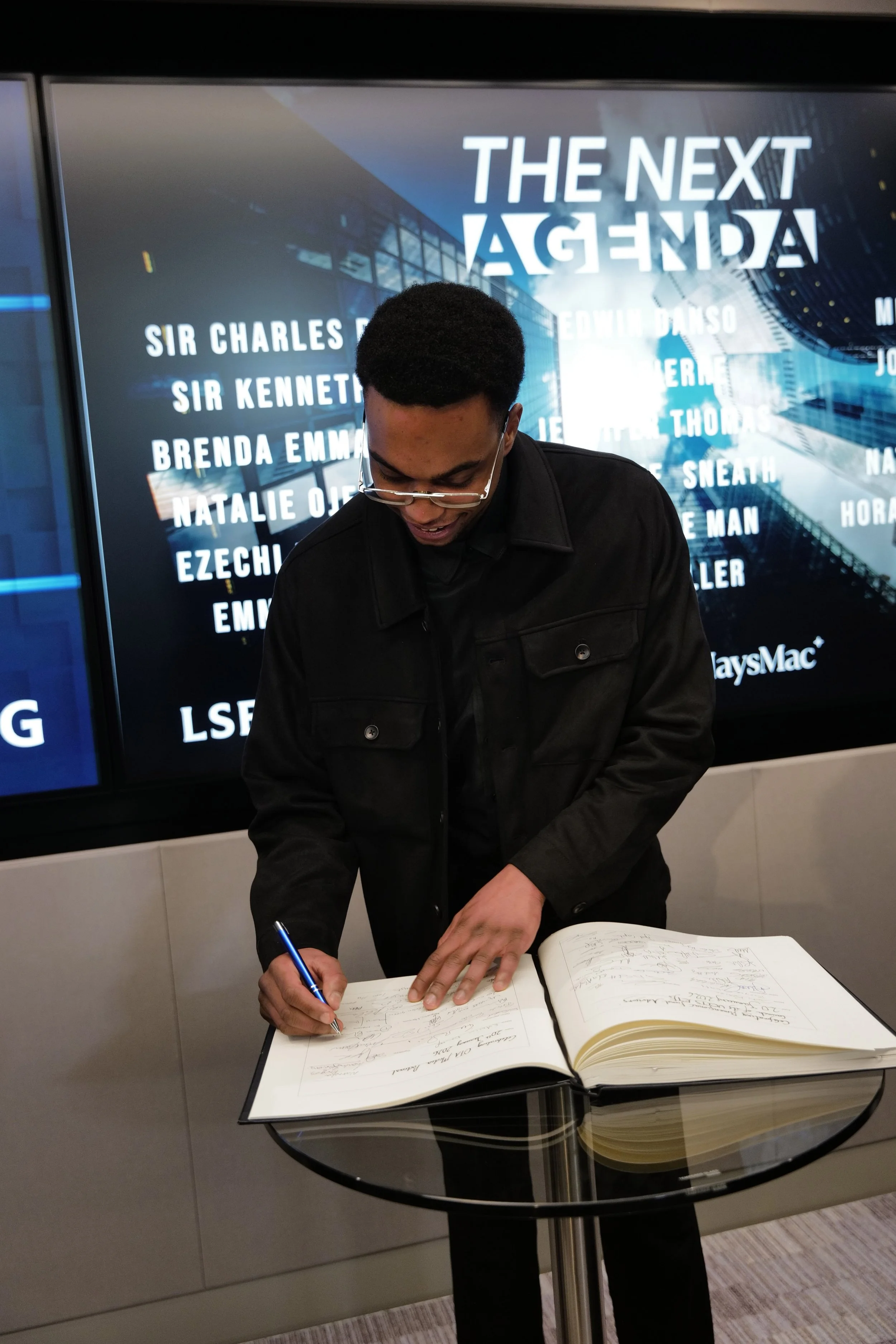 A man in glasses and a black jacket signs a large book on a glass table in front of a screen displaying 'THE NEXT AGENDA' and multiple names.