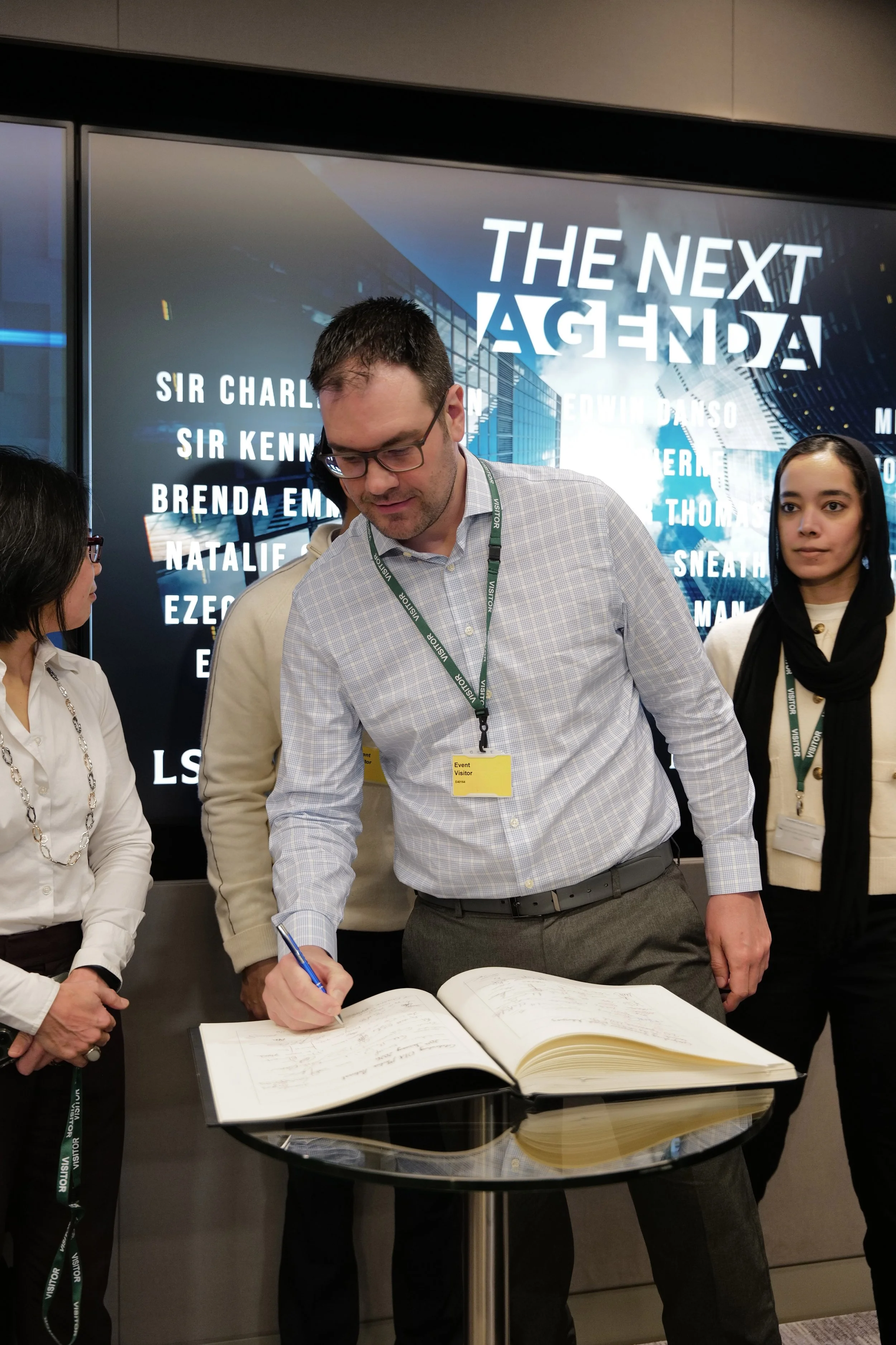 Man with glasses and visitor badge signing a large book on a glass table, with three people standing nearby and a screen behind reading 'The Next Agenda'.