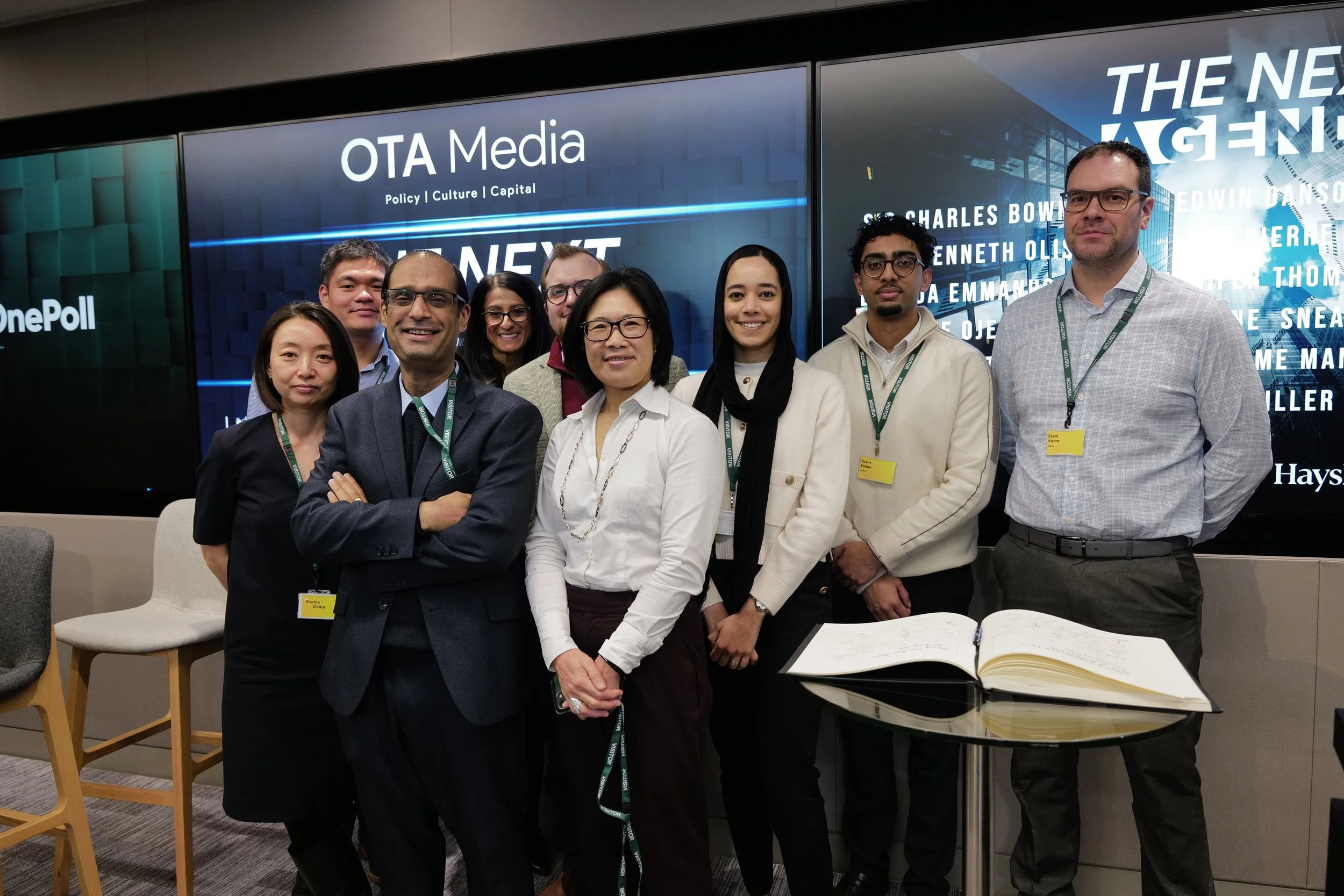 Group of eight diverse professionals standing in front of screens displaying 'OTA Media' and other text, with a glass table and an open book in the foreground.