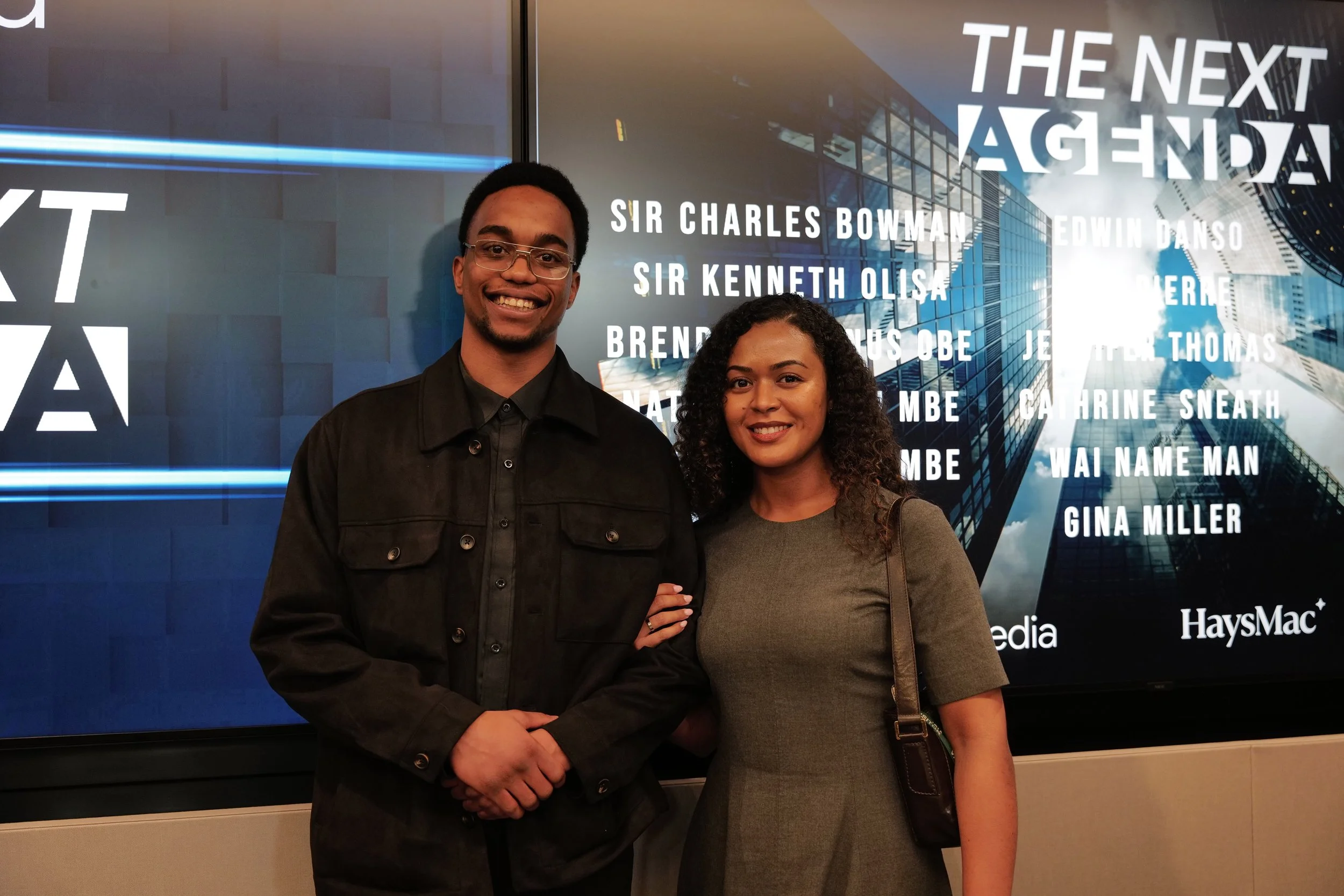 Smiling man and woman standing arm in arm in front of a screen displaying 'THE NEXT AGENDA' and a list of names.