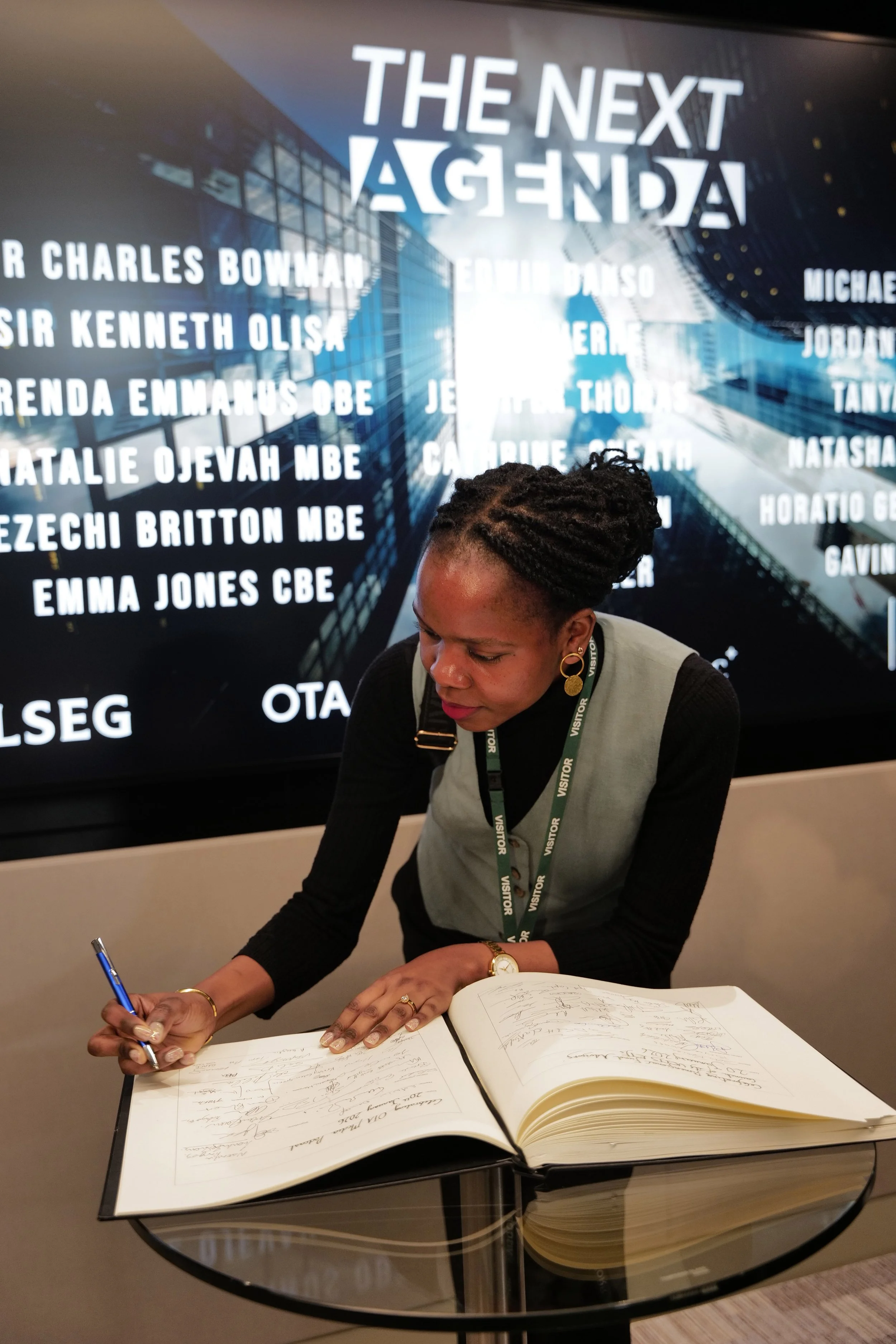 Woman with braided hair wearing a visitor badge signs a large guest book on a glass table, with a digital display behind showing the text 'THE NEXT AGENDA' and a list of names.