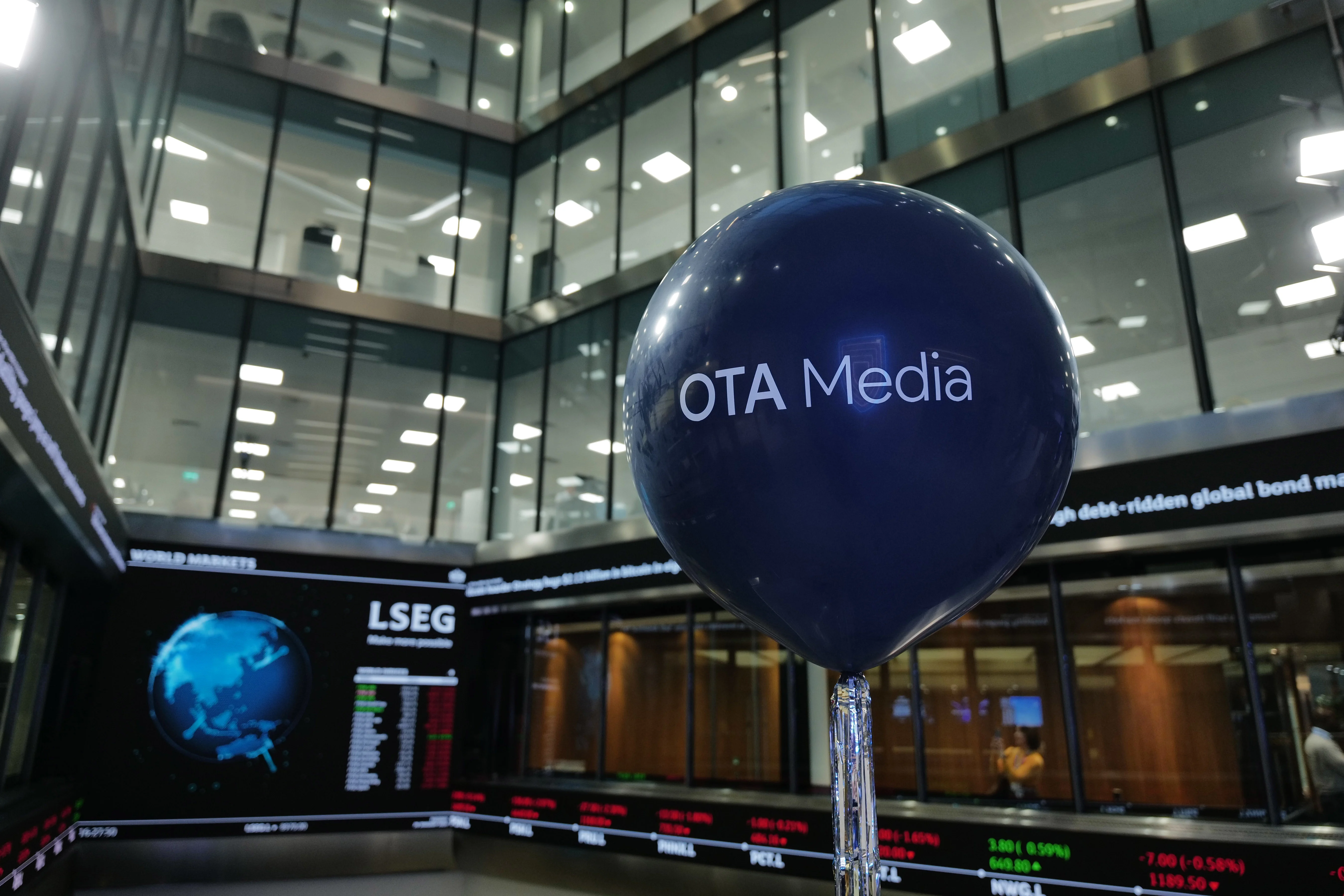 Dark blue balloon with 'OTA Media' text in a modern office building with glass walls and digital stock market displays.