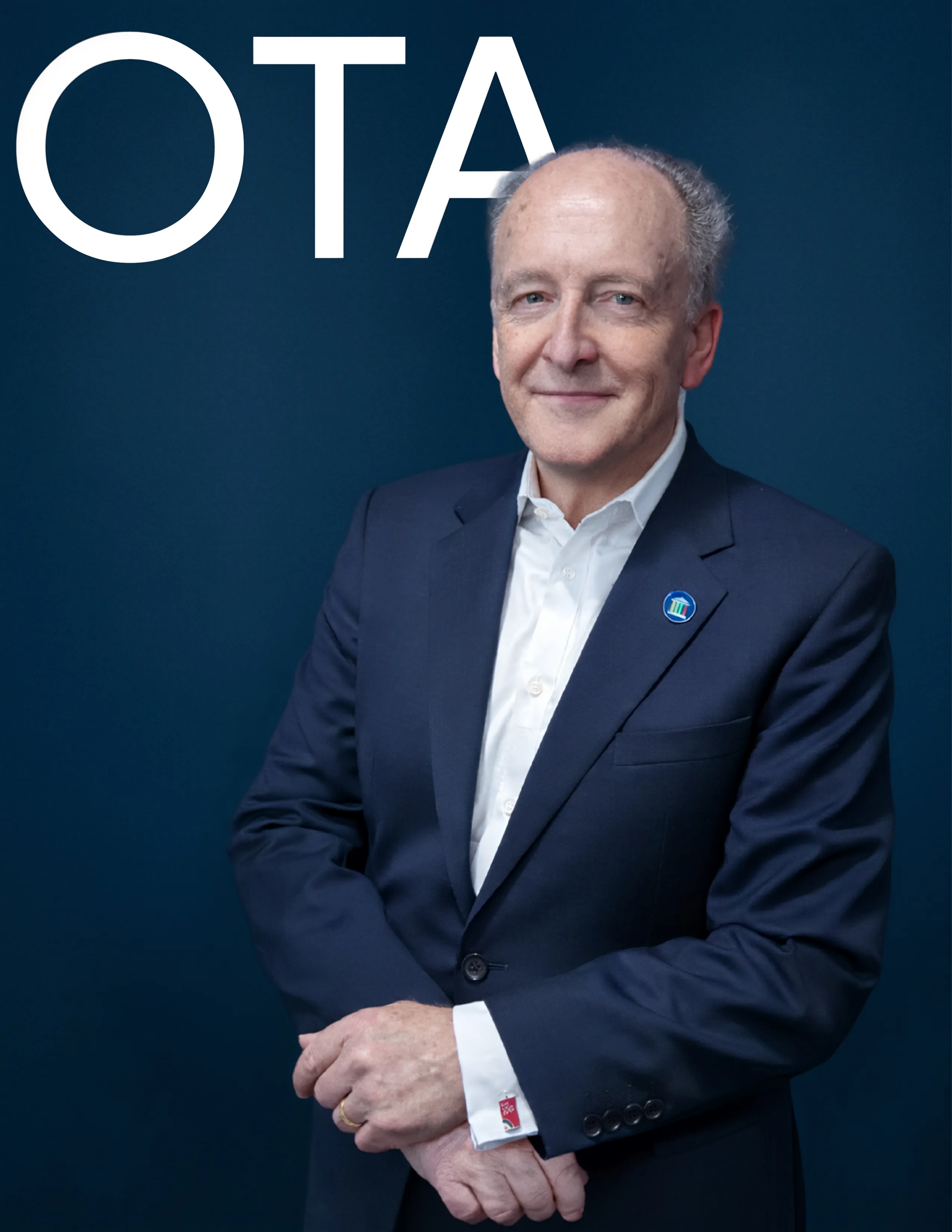 Smiling older man in a navy suit and white shirt standing against a dark blue background with large white letters 'OTA'.