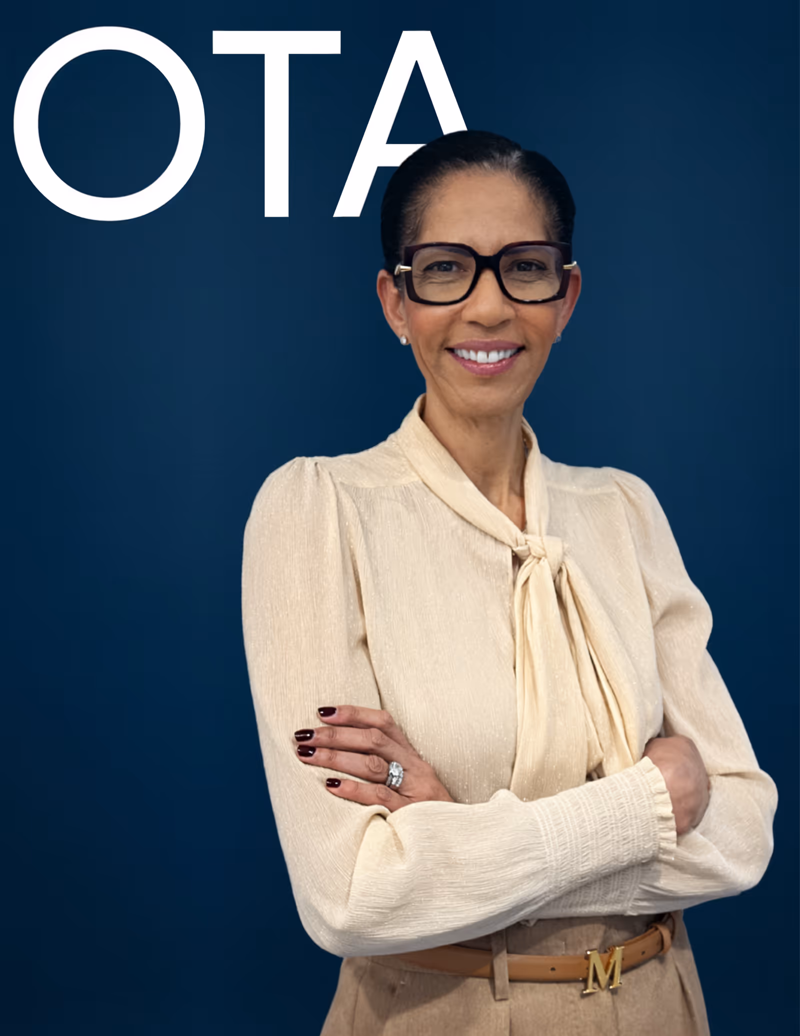 Confident woman with black glasses, cream blouse, and tan pants standing with arms crossed against a dark blue background with large white letters 'OTA'.