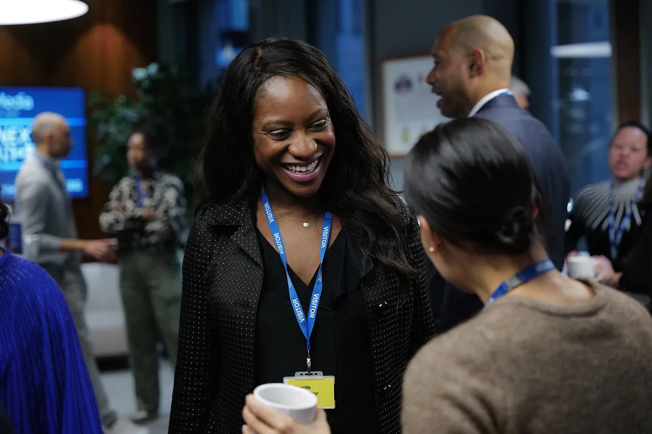 Smiling woman wearing a visitor badge talking to another person holding a cup at a professional event.