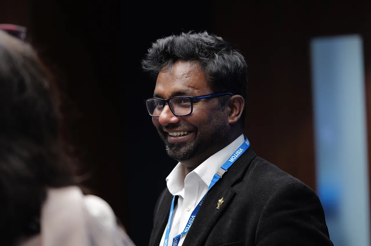 Smiling man with glasses wearing a white shirt, black jacket, and blue visitor badge.
