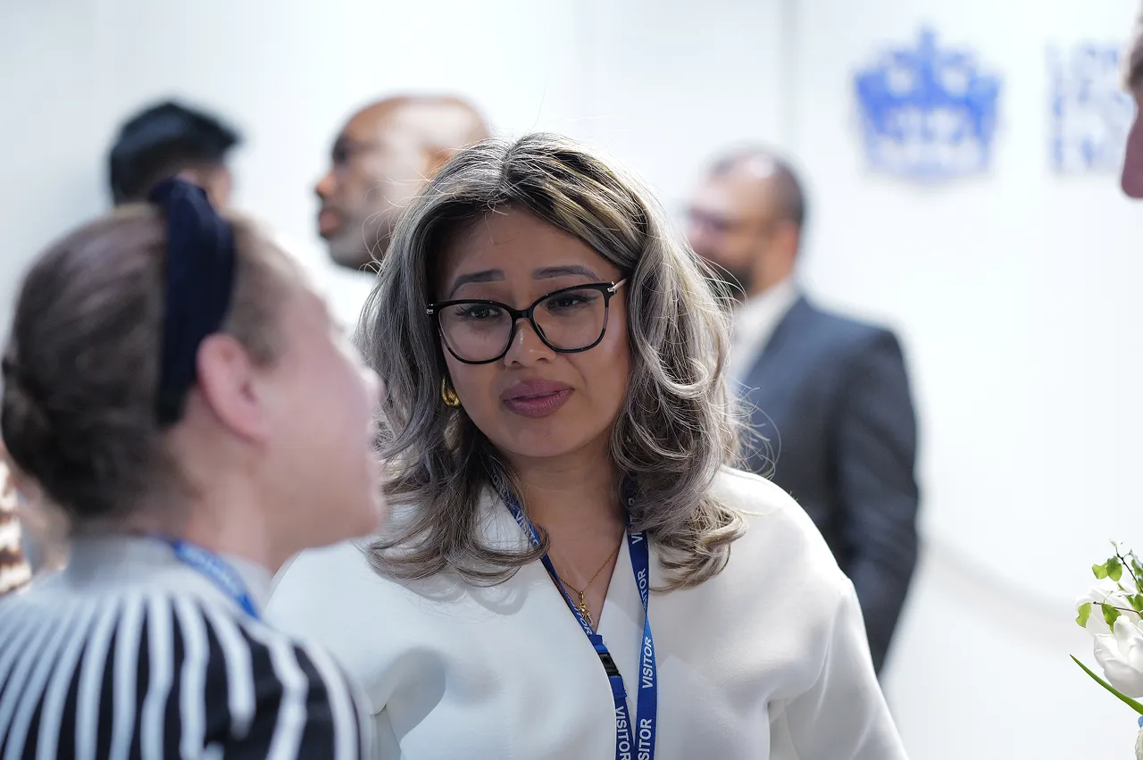 Woman with glasses and gray hair wearing a visitor badge, engaging in conversation at a professional event.