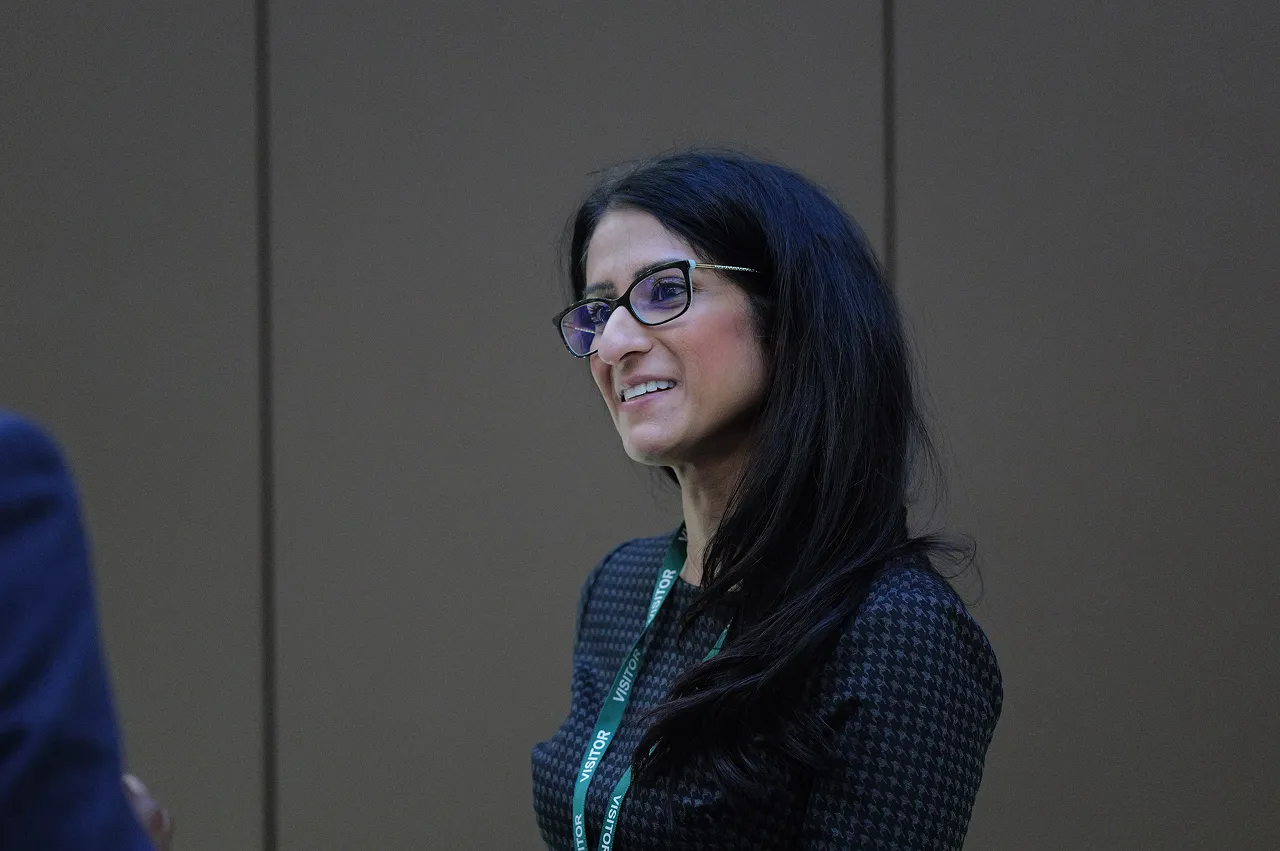 Smiling woman with long dark hair and glasses wearing a dark patterned top and a green visitor lanyard.