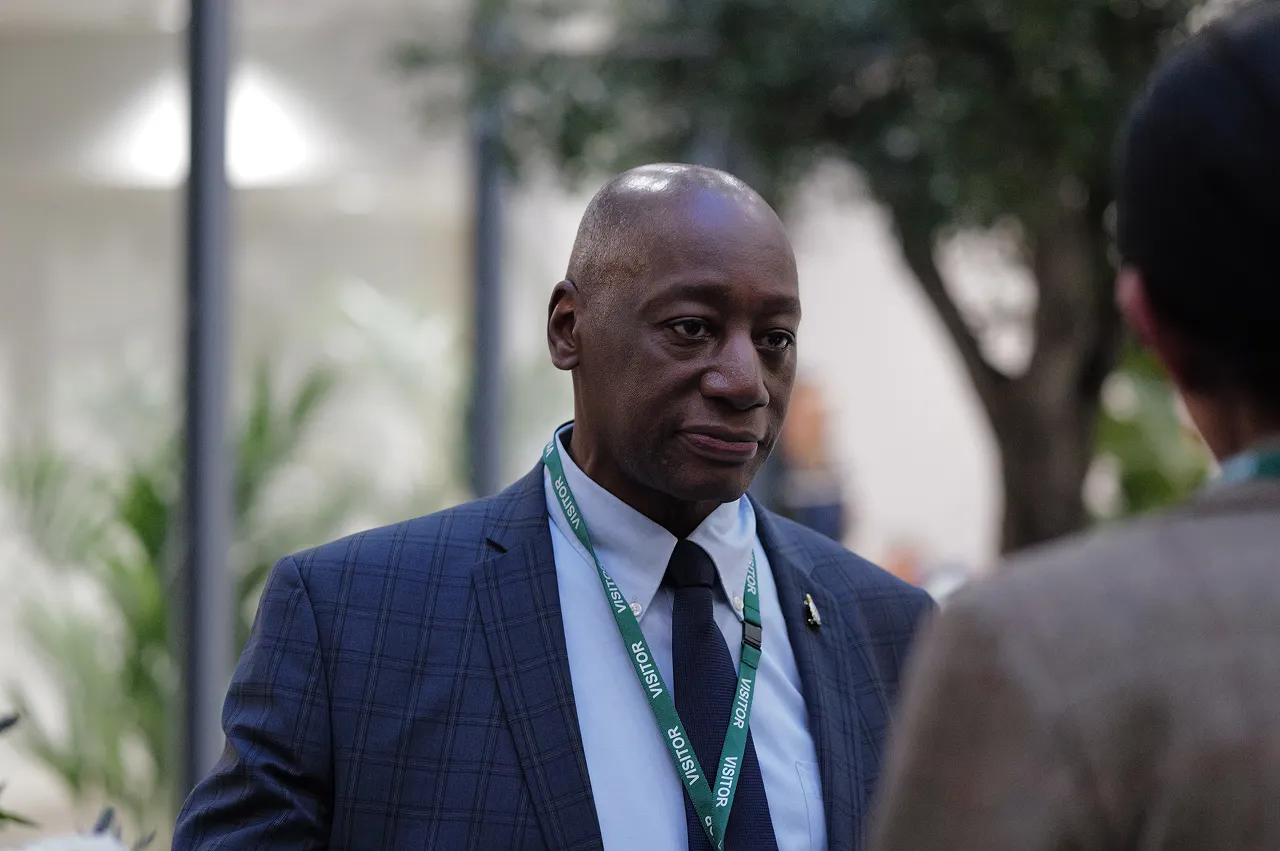 Man in a blue checkered suit and tie wearing a green visitor badge lanyard, engaged in conversation indoors.
