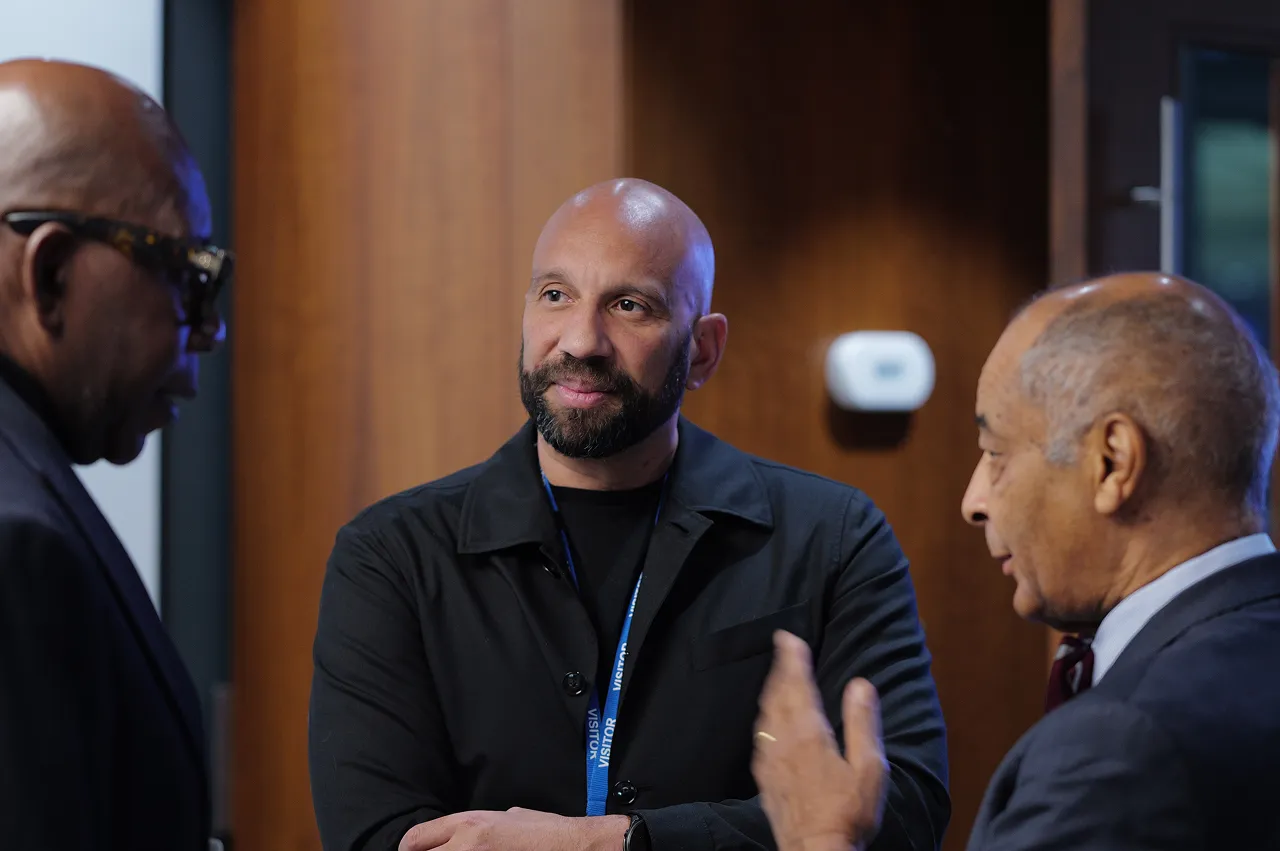 Three men engaged in a conversation in a professional setting, one man in the center listens attentively with arms crossed.