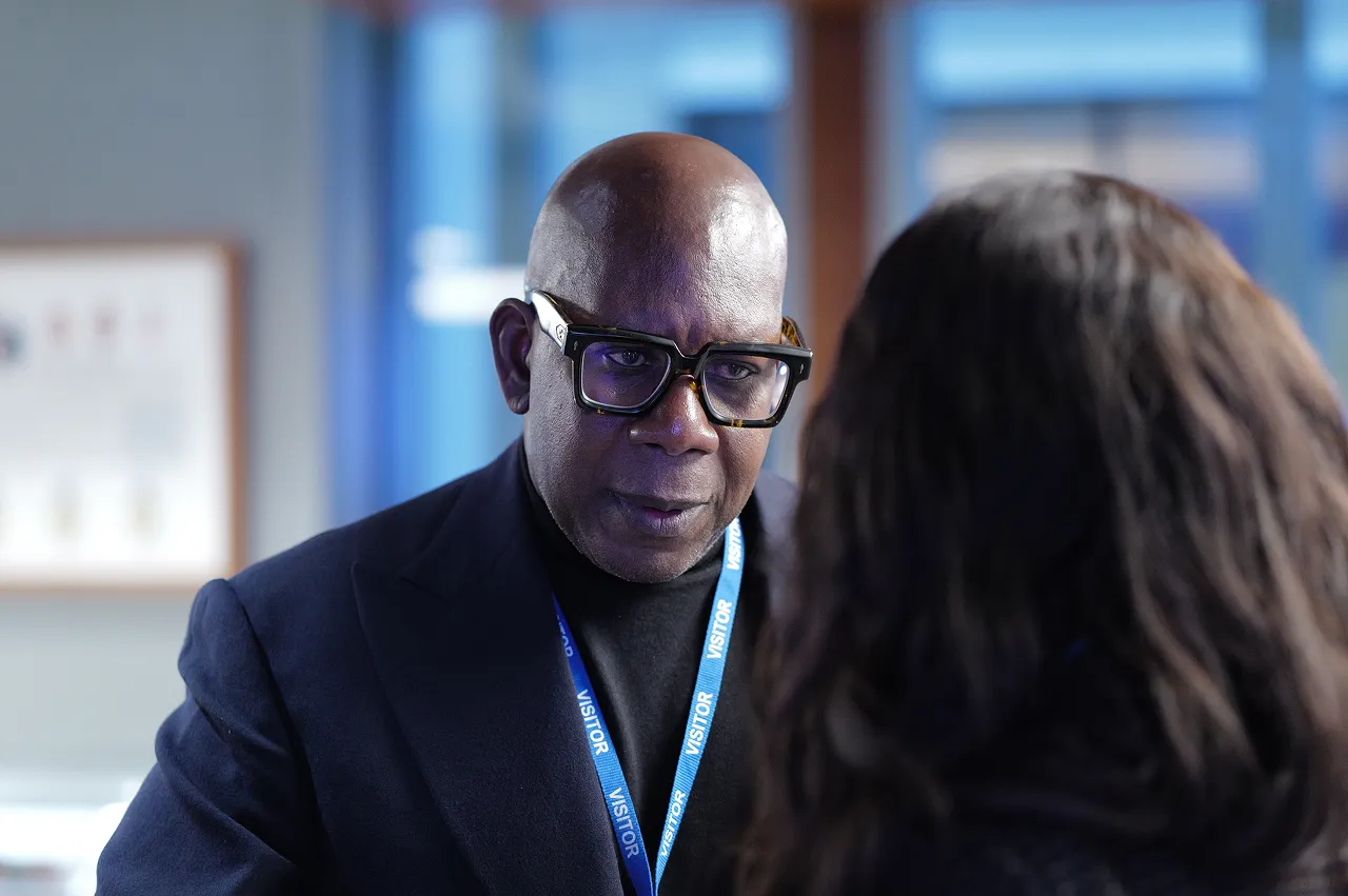 Man wearing large glasses and a visitor badge, engaged in serious conversation with a woman whose back is to the camera.