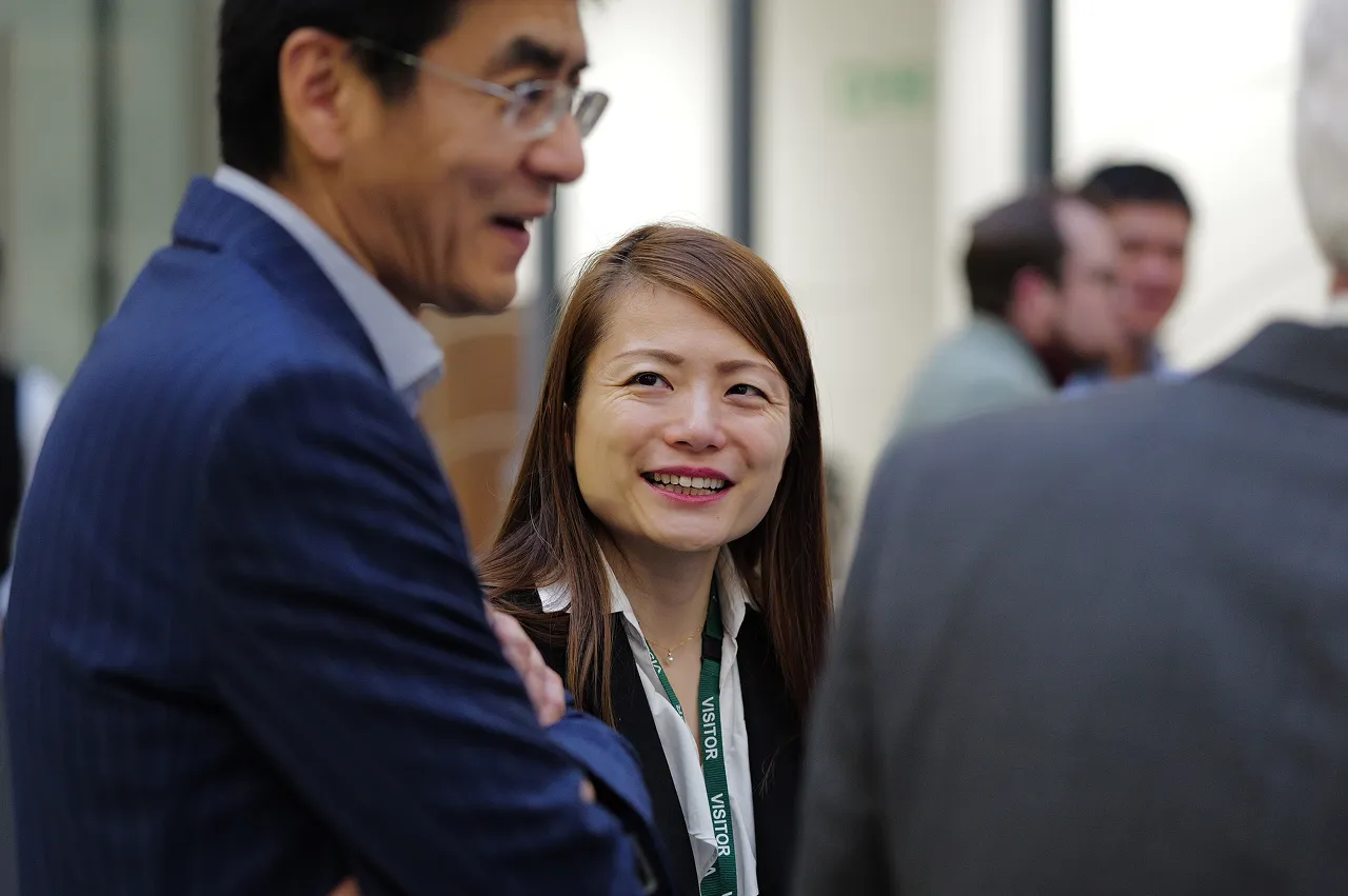 Woman with a visitor badge smiling and talking with people at a professional event.