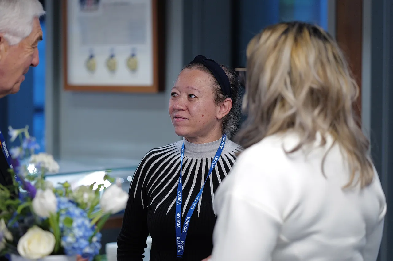 Three people engaged in conversation indoors, one woman wearing a visitor badge and a black headband.