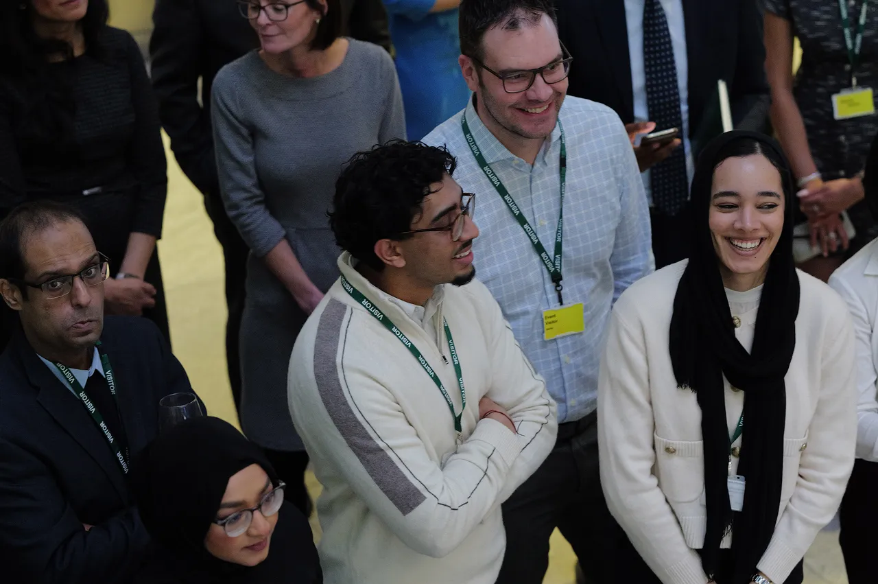 A diverse group of people wearing visitor badges standing indoors, some smiling and engaging in conversation.