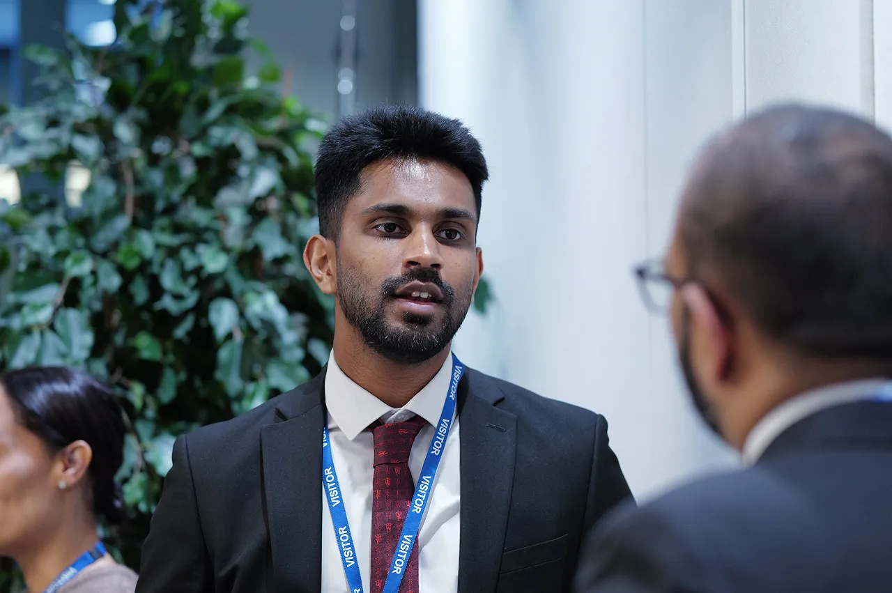 Man in a black suit and red tie wearing a blue visitor lanyard speaking to another person indoors.