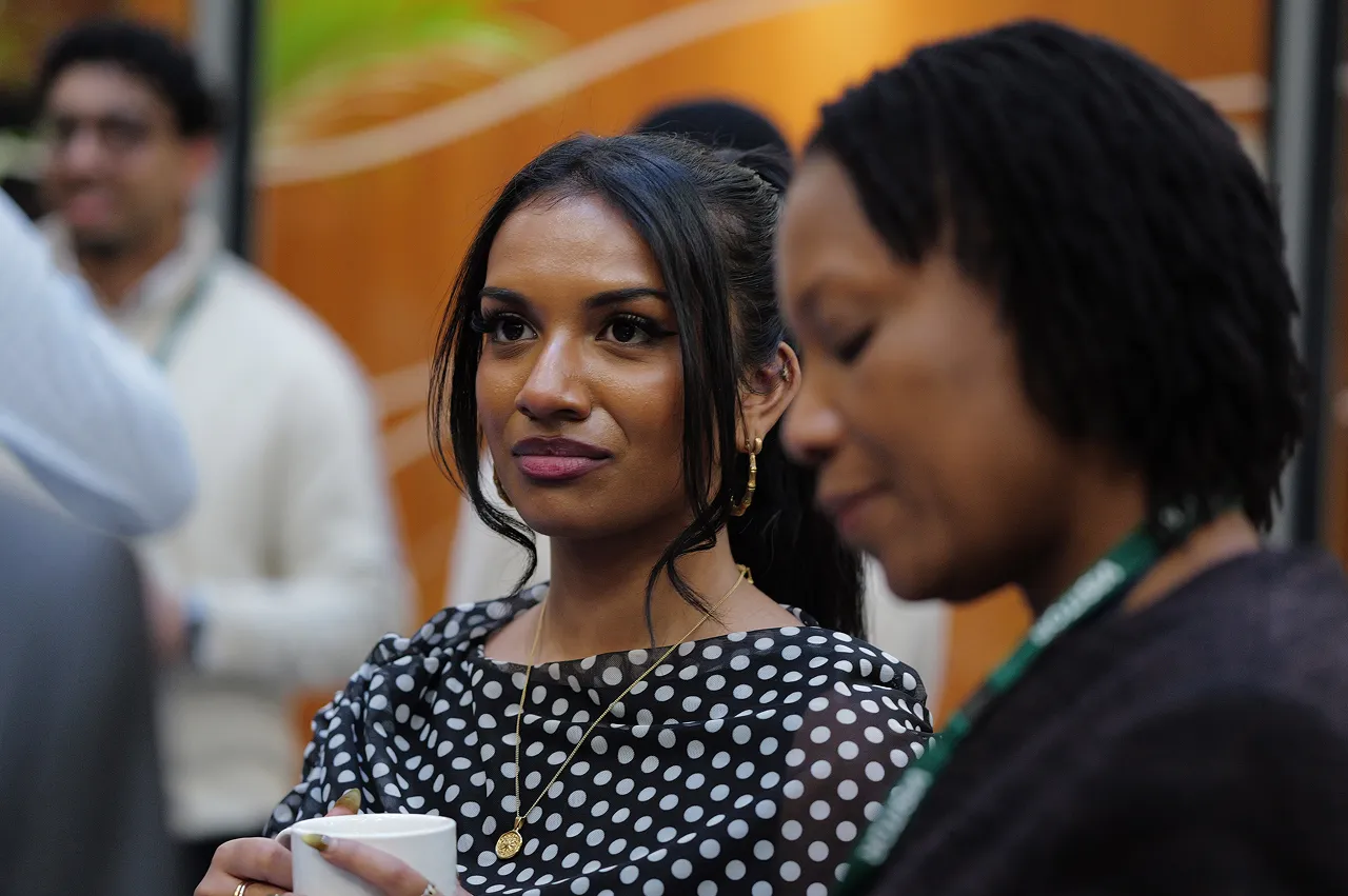 Two women at an event, one holding a white cup and wearing a black and white polka dot top, the other looking down with a green lanyard.
