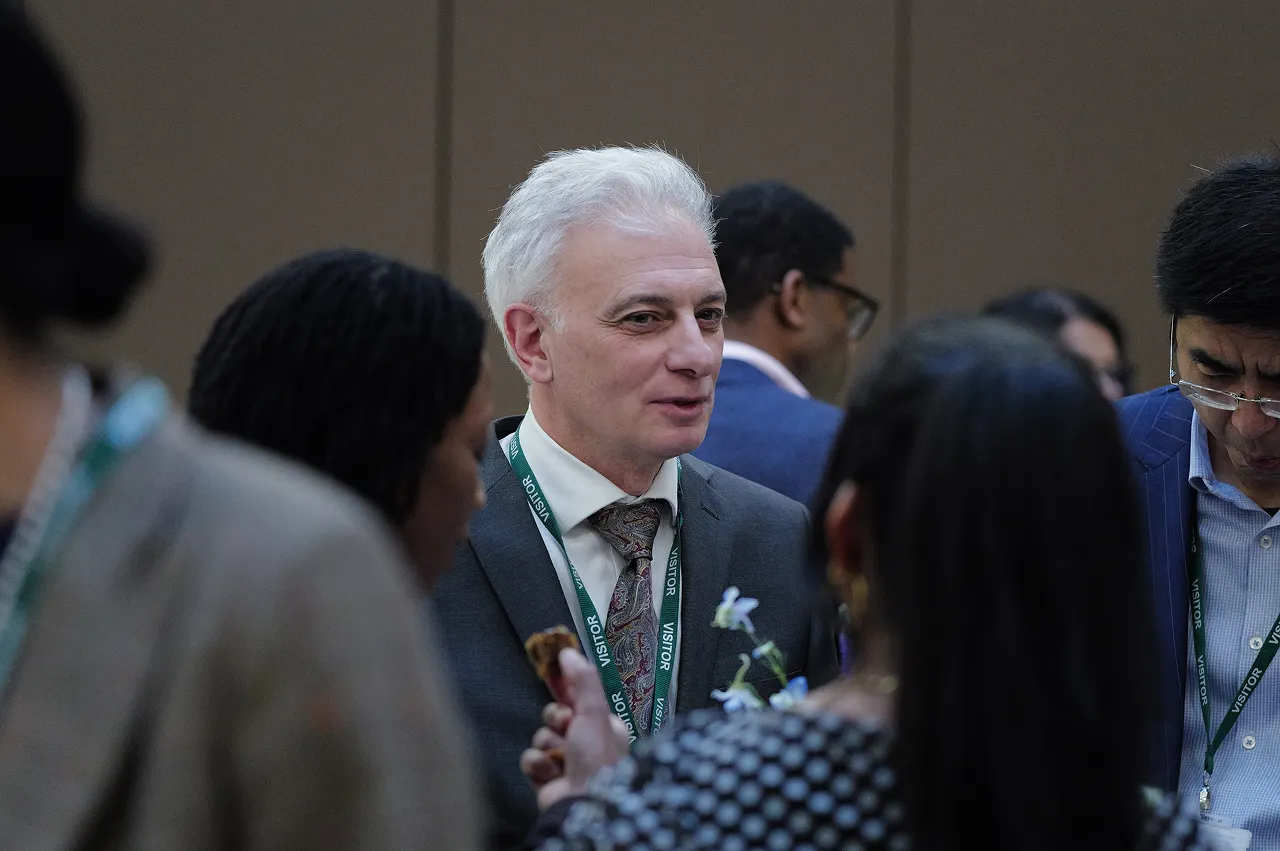 Man in a suit with white hair and visitor badge talking with a group of people at an indoor event.