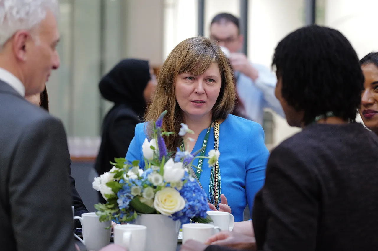 Group of people engaged in conversation around a table with white and blue flower centerpiece and white coffee cups.