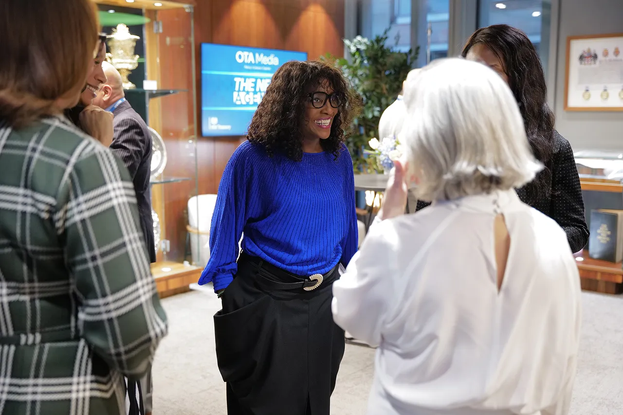 A group of people engaged in conversation, featuring a woman in a bright blue sweater smiling and interacting with others in a professional setting.