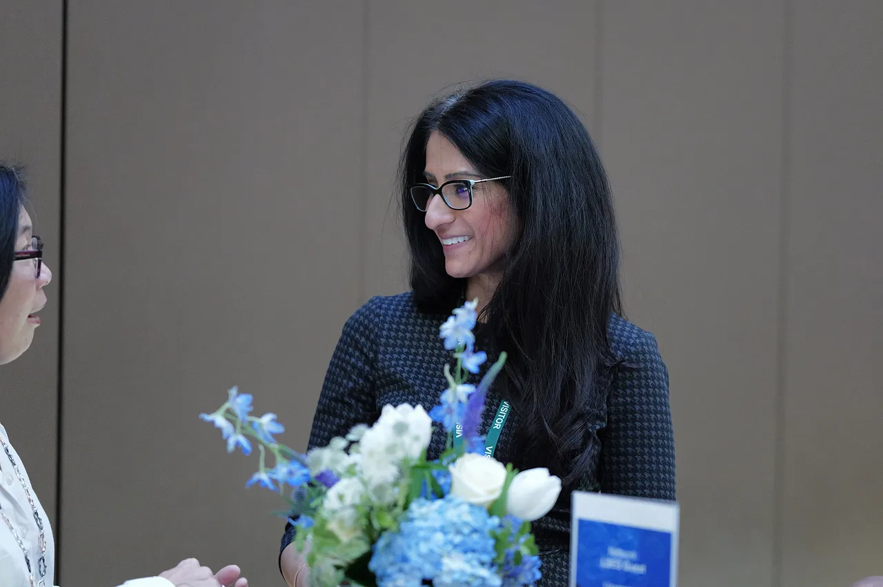Two women talking, one holding a bouquet of blue and white flowers, both wearing glasses.