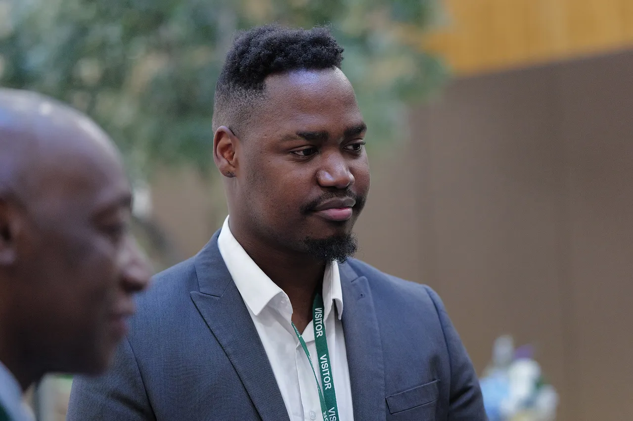 Man in a gray suit and white shirt wearing a green visitor badge standing indoors, with another blurred man in the foreground.