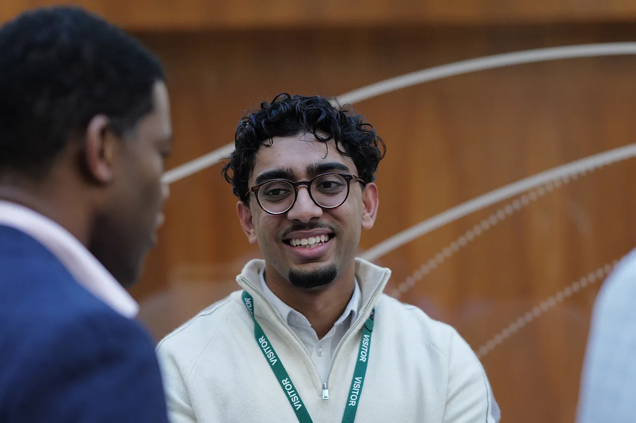 Smiling young man with glasses wearing a white zip-up sweater and visitor badge talking to another person.