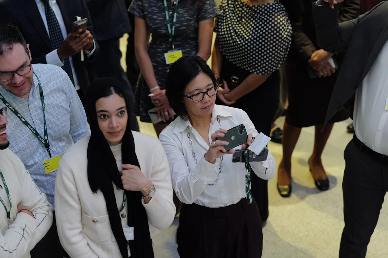 Group of diverse people standing indoors, some looking upward and one woman in glasses taking a photo with her smartphone.