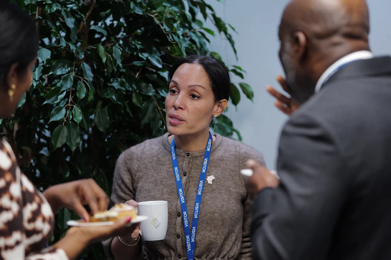 Three professionals engaged in conversation during a break, one woman holding a cup and another holding a plate with food.