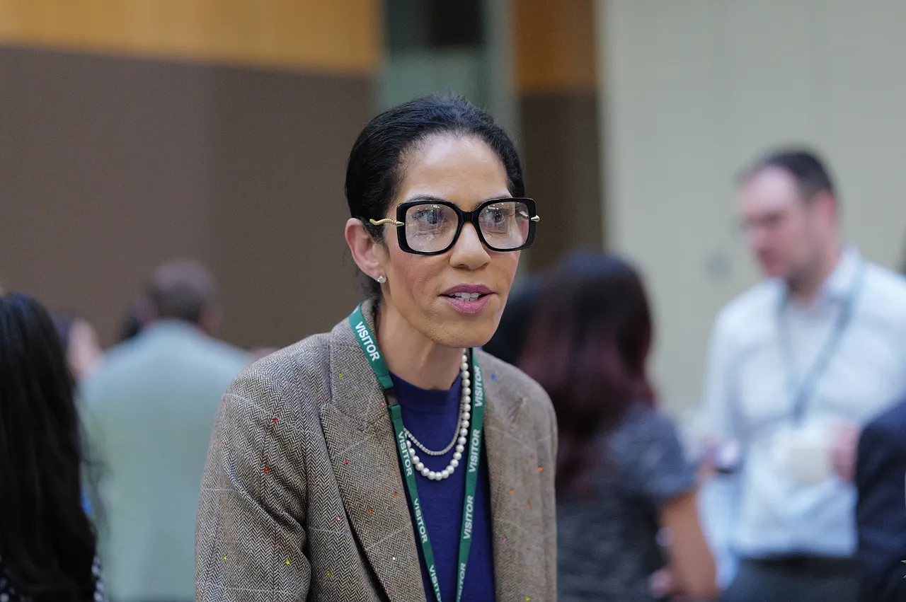 Woman with dark hair tied back wearing large black glasses, a brown blazer, and a visitor badge, speaking indoors with people blurred in the background.