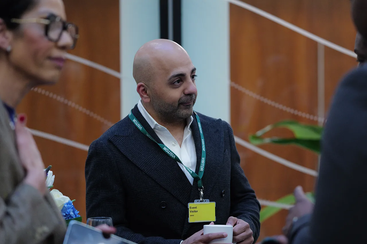 Man wearing a visitor badge and holding a white cup, standing indoors and listening attentively.