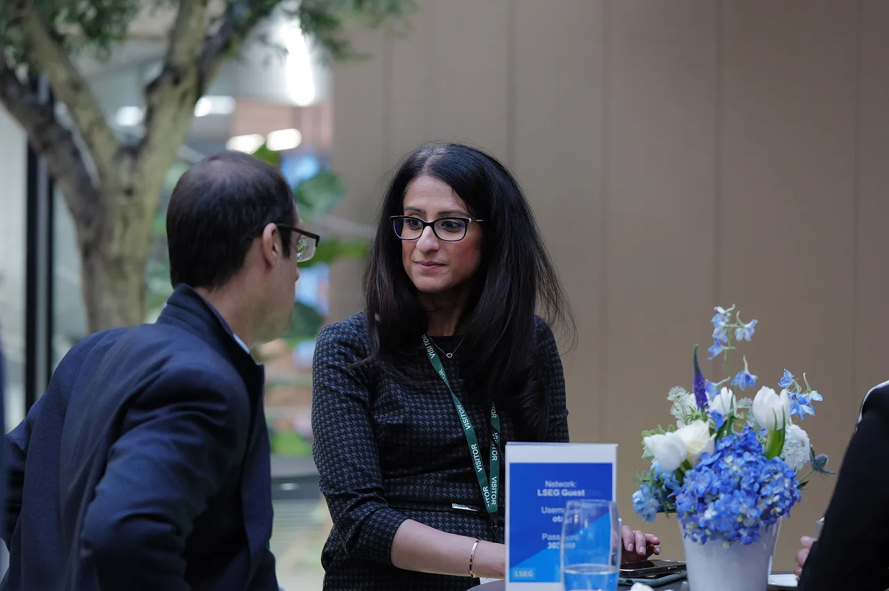 Two professionals wearing glasses engaged in conversation at a table with a blue flower arrangement and a network sign.