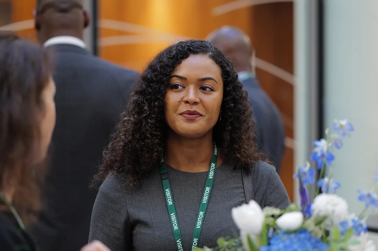 Woman with curly hair wearing a grey top and green visitor badge, standing near a bouquet of blue and white flowers.