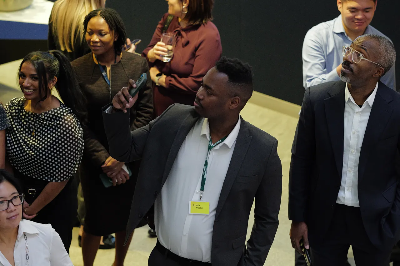 A diverse group of people at an event, including a man in a suit taking a selfie with a smartphone.