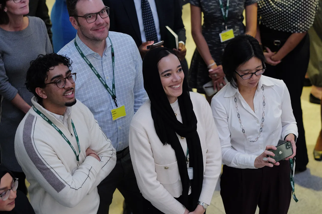 A diverse group of young adults, some wearing visitor badges, attentively watching an event, with one woman holding a smartphone.