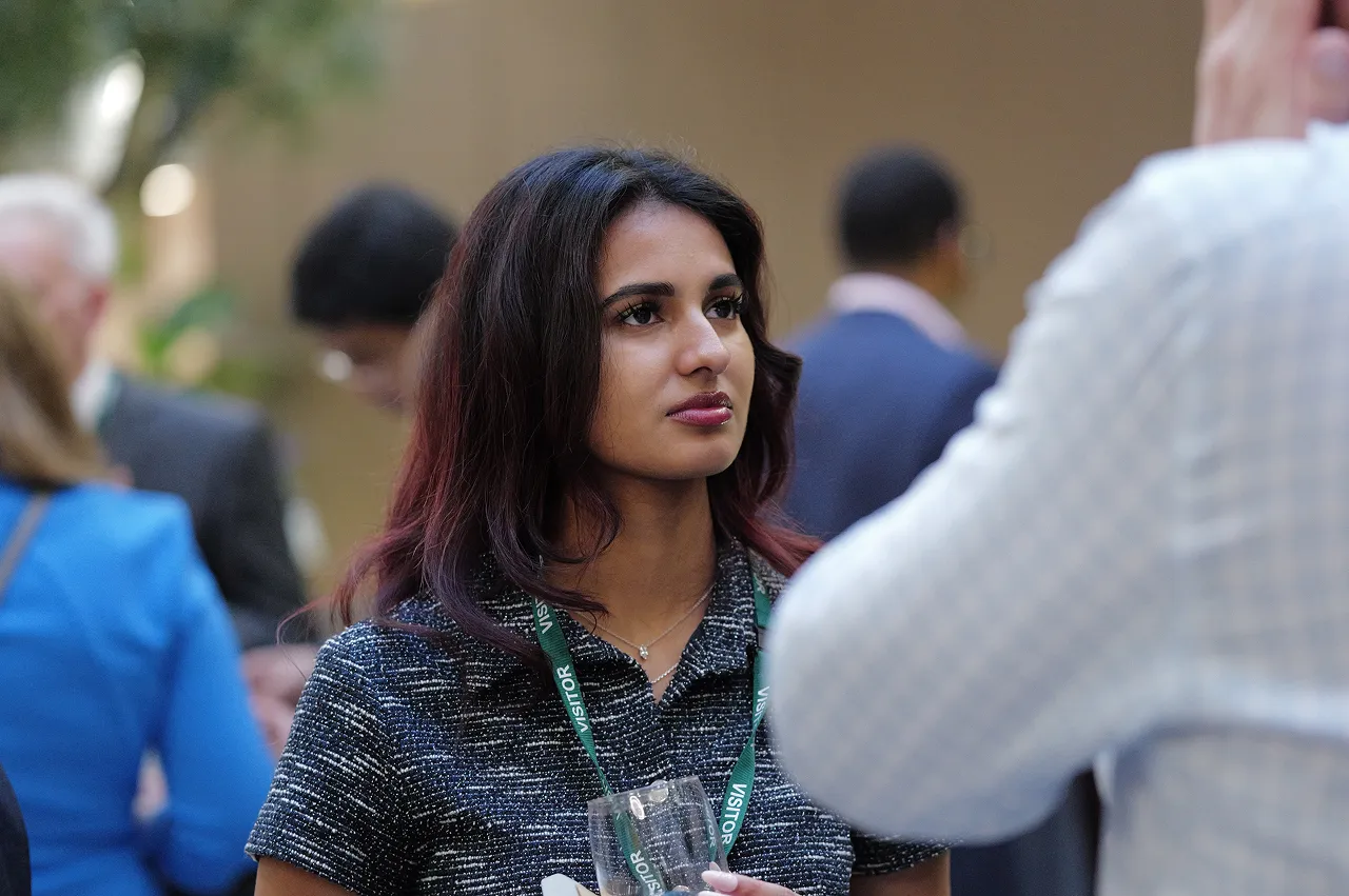 Young woman with dark hair wearing a visitor badge, listening attentively in a busy indoor setting.