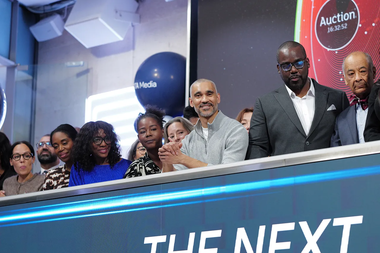 A diverse group of people standing behind a counter with digital screens, some smiling, with a balloon and auction timer in the background.