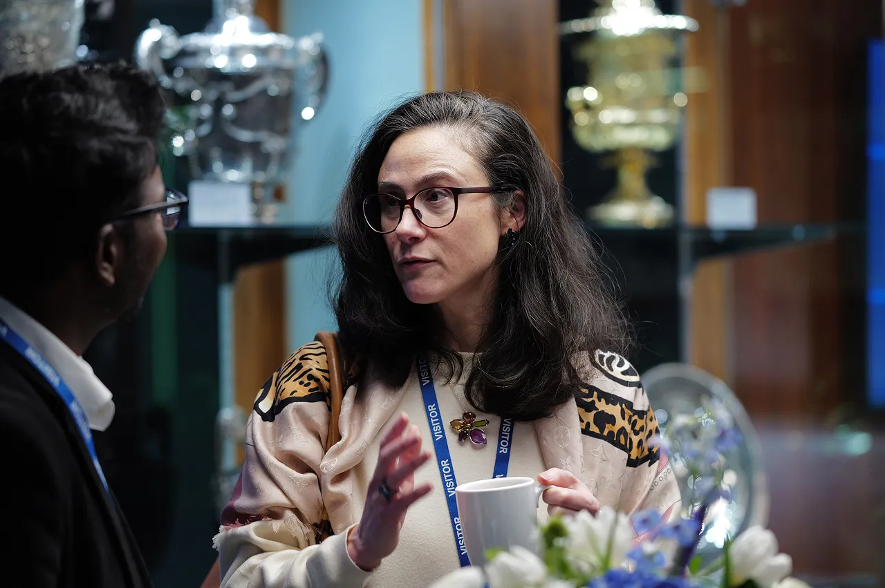 Woman wearing glasses and a visitor badge holding a mug and talking to a man in an indoor setting with trophies in the background.
