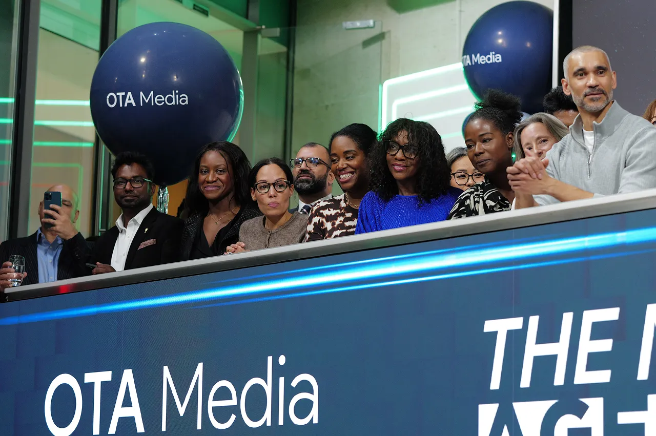 A diverse group of people standing behind a display with the OTA Media logo, smiling and clapping at an event.
