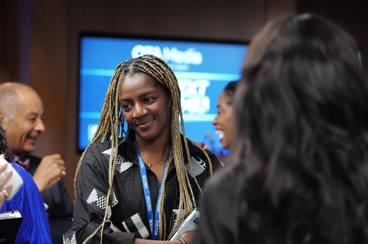 A woman with braided hair wearing a visitor badge, standing with arms crossed and smiling thoughtfully in a social gathering.