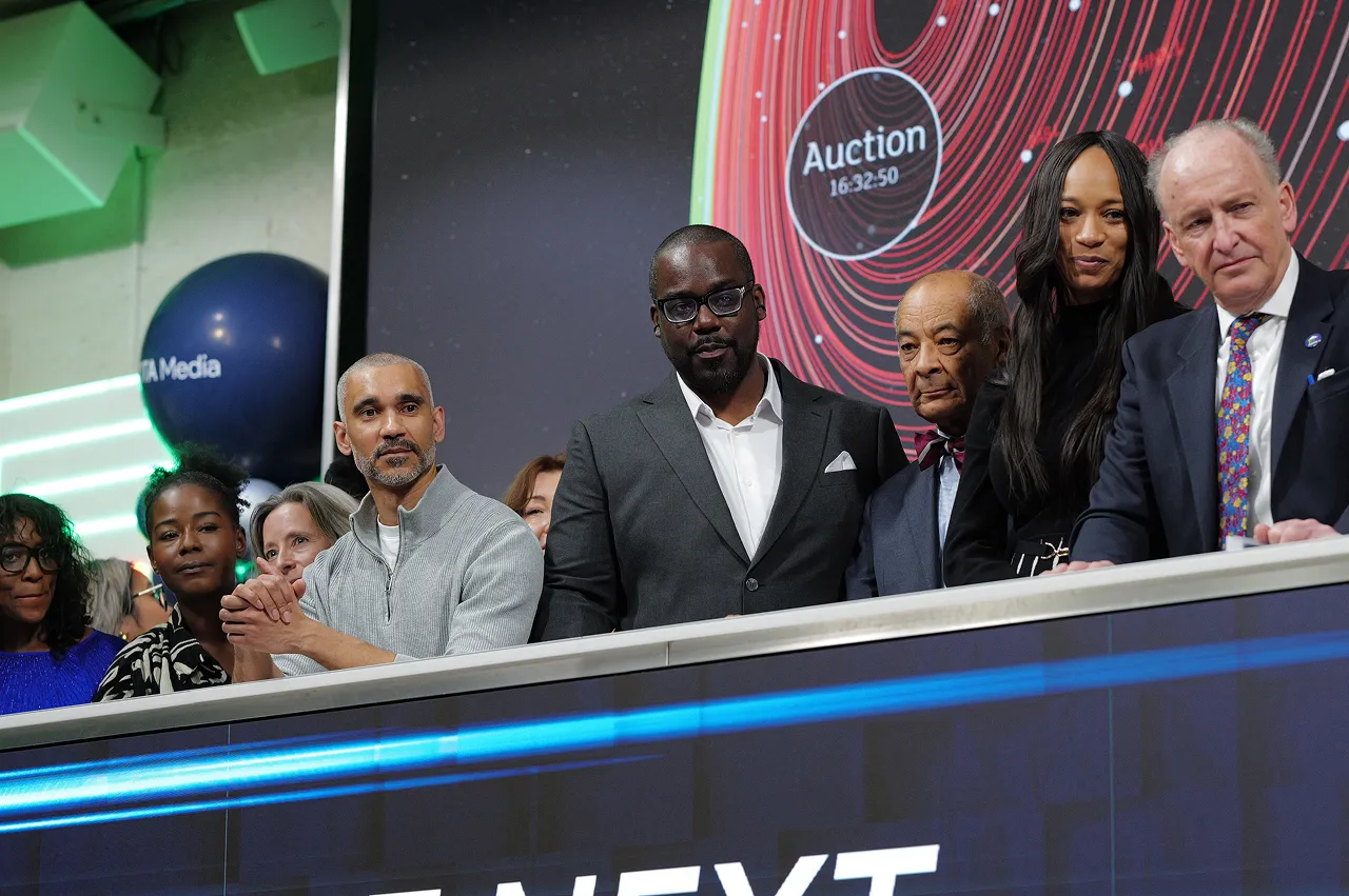 A group of seven people standing behind a podium with a digital display showing the word 'Auction' and a countdown timer in the background.