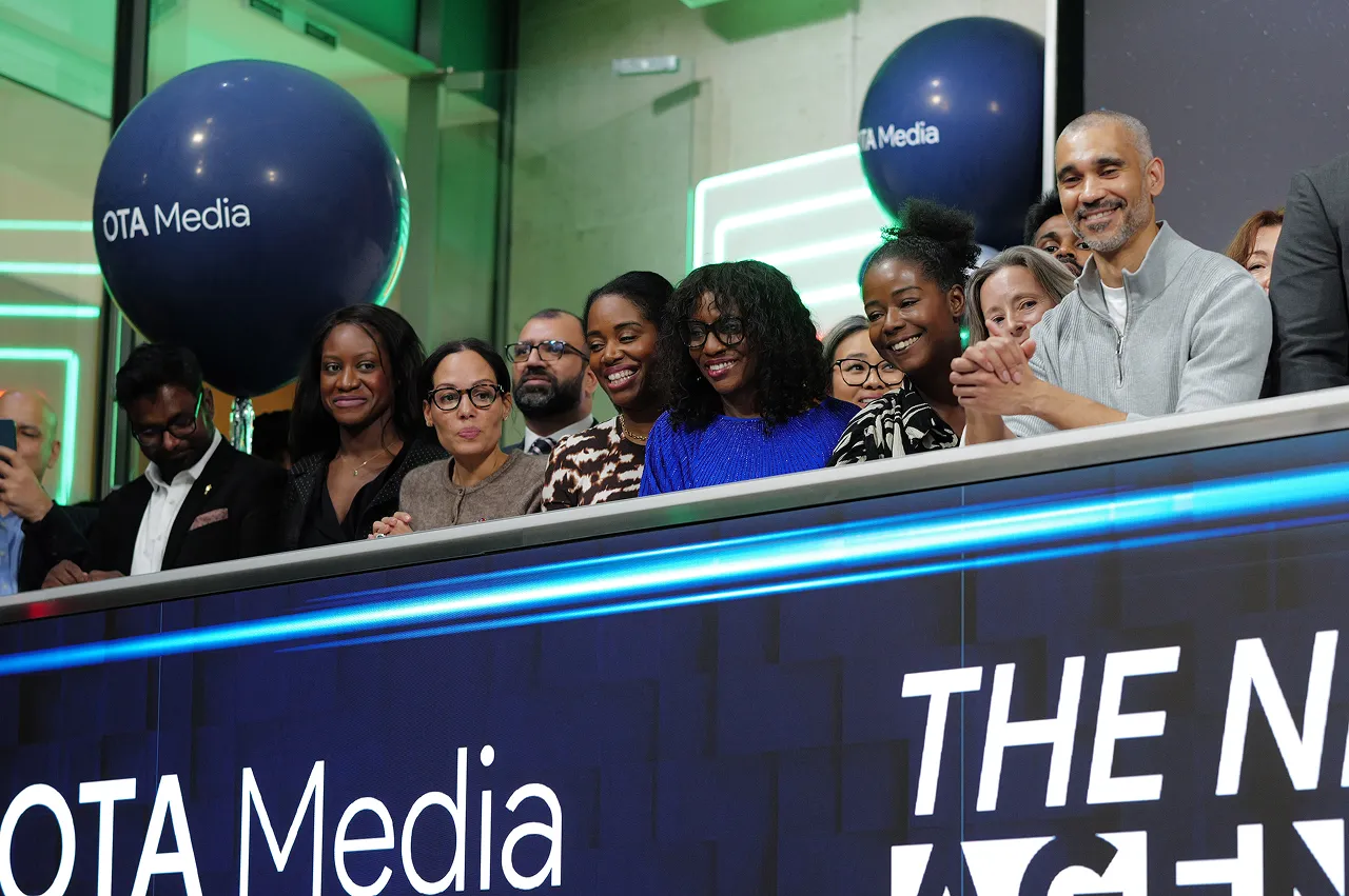 A diverse group of people smiling and standing behind a digital board with 'OTA Media' balloons in the background.