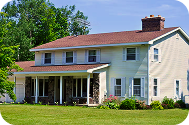 Two-story house with a red roof, light yellow siding, a covered front porch, and green bushes in front under a clear blue sky.
