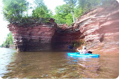 Person kayaking near a large red rock cliff and tree-covered shoreline in clear water.