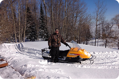 Man standing next to a yellow snowmobile on a snowy path surrounded by leafless trees.